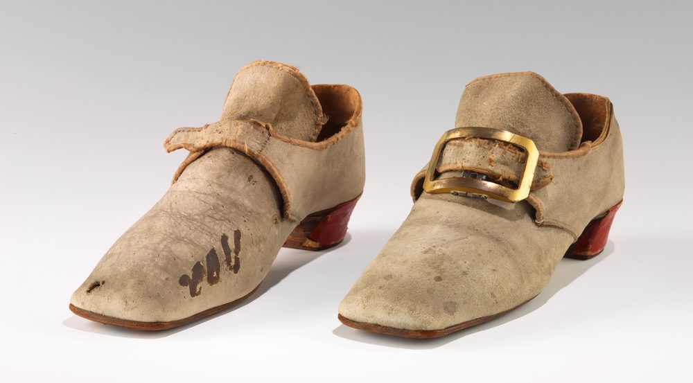Par de zapatos de ante claro con punta cuadrada, hebilla dorada y tacón rojo.